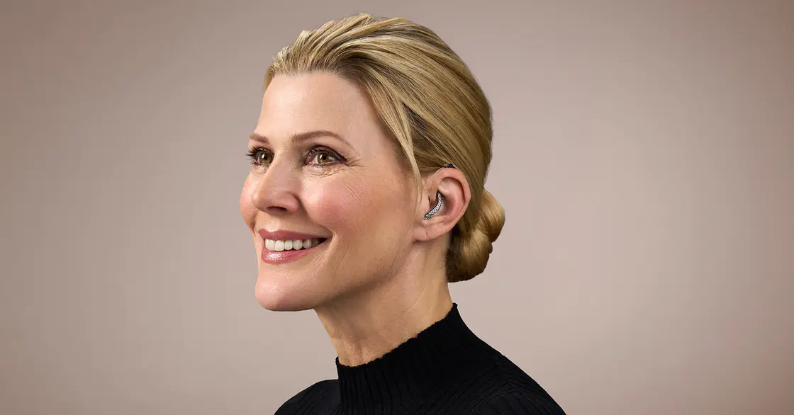 Lächelnde Frau mit blondem Haar in einem tiefen Dutt, mit schwarzem Oberteil und Hörgerät, vor einem neutralen Hintergrund.