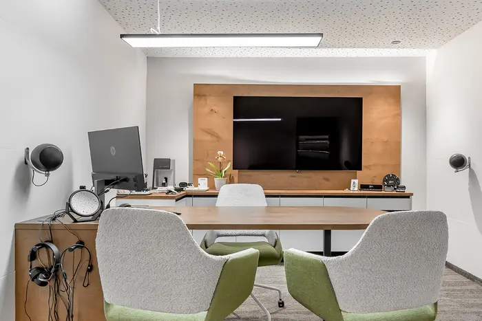 Modernes Büro mit zwei grauen Stühlen, einem Holzschreibtisch, einem Computer und einem großen, an der Wand befestigten Bildschirm gegenüber den Stühlen.