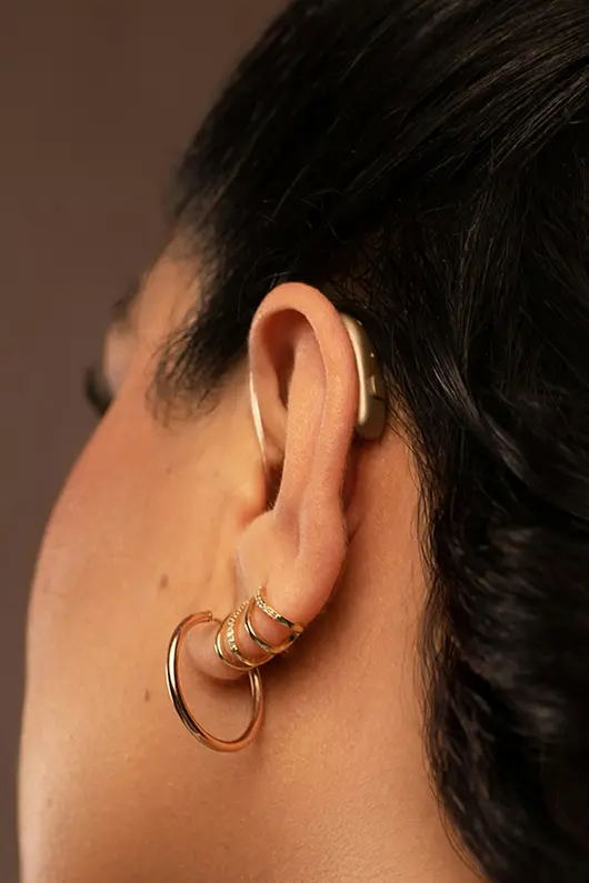 Nahaufnahme von dem Ohr einer Frau. Sie trägt ein Hinter-dem-Ohr-Hörgerät und goldene Ohrringe.