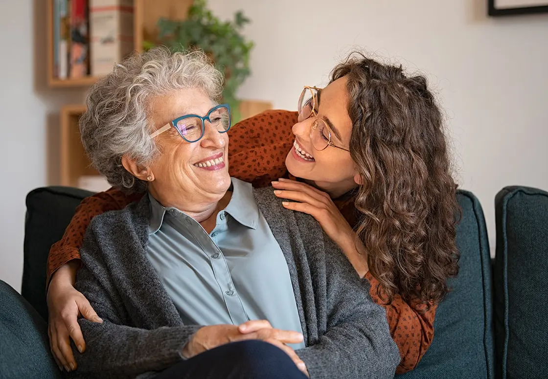 Ältere Frau sitzt auf einer Couch, eine jüngere Frau umarmt sie von hinten und beide lächeln und umarmen sich herzlich.
