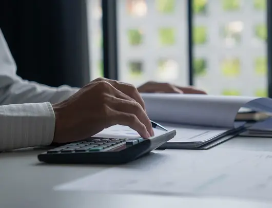 Person, die einen Taschenrechner benutzt und in einem Notizbuch an einem Schreibtisch in der Nähe eines Fensters mit Dokumenten und einem Stifthalter schreibt.