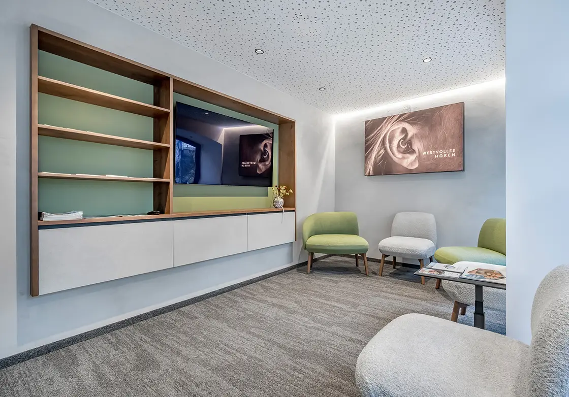 Modernes Wartezimmer mit grünen Stühlen, einem an der Wand befestigten Regal und gerahmten Foto eines Ohrs an der Wand.