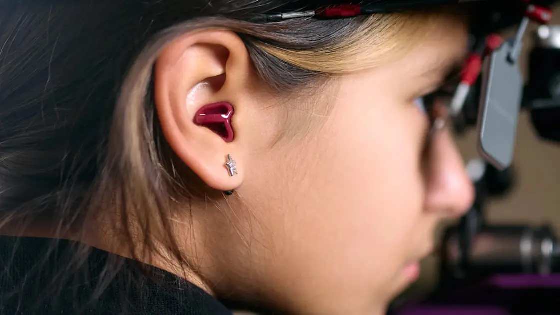 Nahaufnahme des Seitenprofils eines Mädchens, welches einen Ohrstöpsel als Gehörschutz im Ohr trägt, mit kurzen Haaren und einem dunklen Hintergrund.
