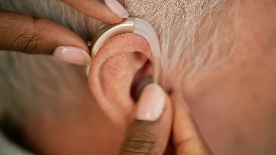 Nahaufnahme einer Person, die einem älteren Menschen ein Hörgerät hinter dem Ohr anpasst.