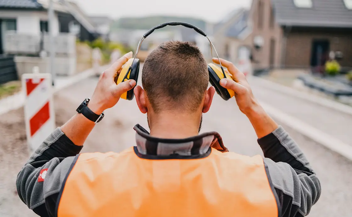 Ein Bauarbeiter in einer orangefarbenen Weste setzt einen Gehörschutz auf, während er auf einer Straße in der Nähe von Gebäuden steht.