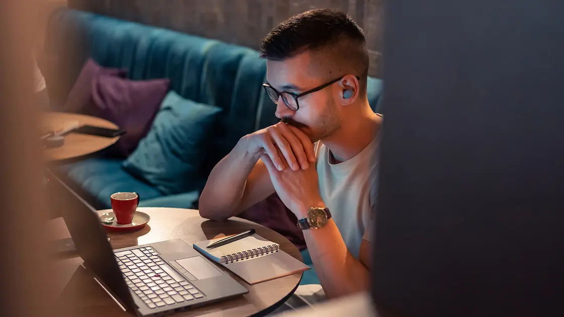 Mann, der an einem Tisch sitzt, auf einen Laptop mit Papieren schaut und eine Tasse Kaffee vor sich stehen hat. Er nimmt an einem online Arbeitsmeeting teil.