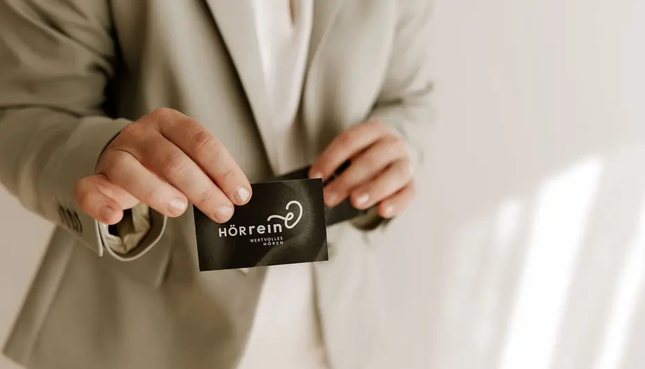 Geschäftsführerin im hellen Anzug, die ihre schwarze Visitenkarte hält, mit unscharfem neutralem Hintergrund und Sonnenlichtschatten.