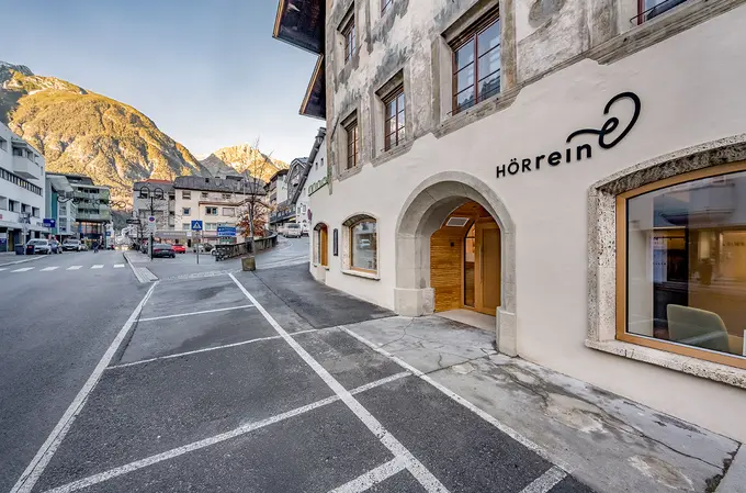 Außenfassade und Umgebung der HÖRrein GmbH in Landeck. Eine ruhige Straße mit leeren Parkplätzen, daneben ein helles Gebäude mit großen Fenstern und Bergen im Hintergrund.