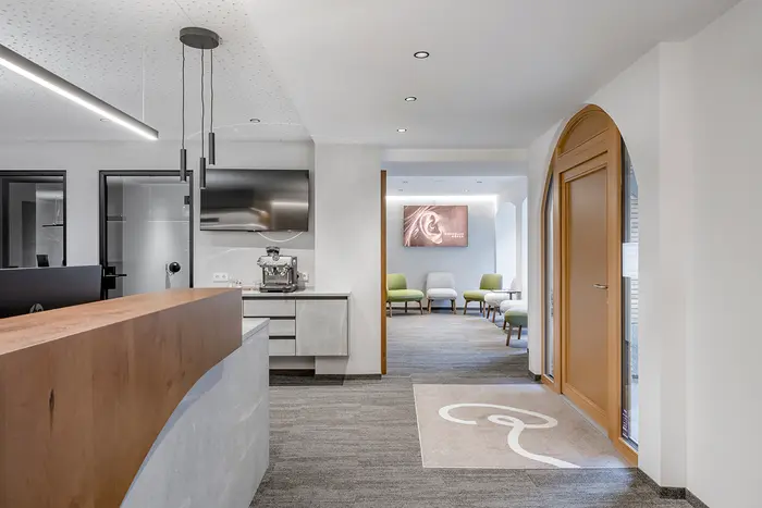 Eingangsbereich der HÖRrein GmbH. Moderner Empfangsbereich mit einem geschwungenen Holzschreibtisch, grauem Teppich und einem Wartezimmer im Hintergrund.