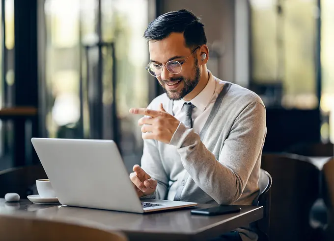 Lächelnder Mann mit Brille, der in einem Café einen Laptop benutzt und auf den Bildschirm zeigt, während er an einem Tisch sitzt. 