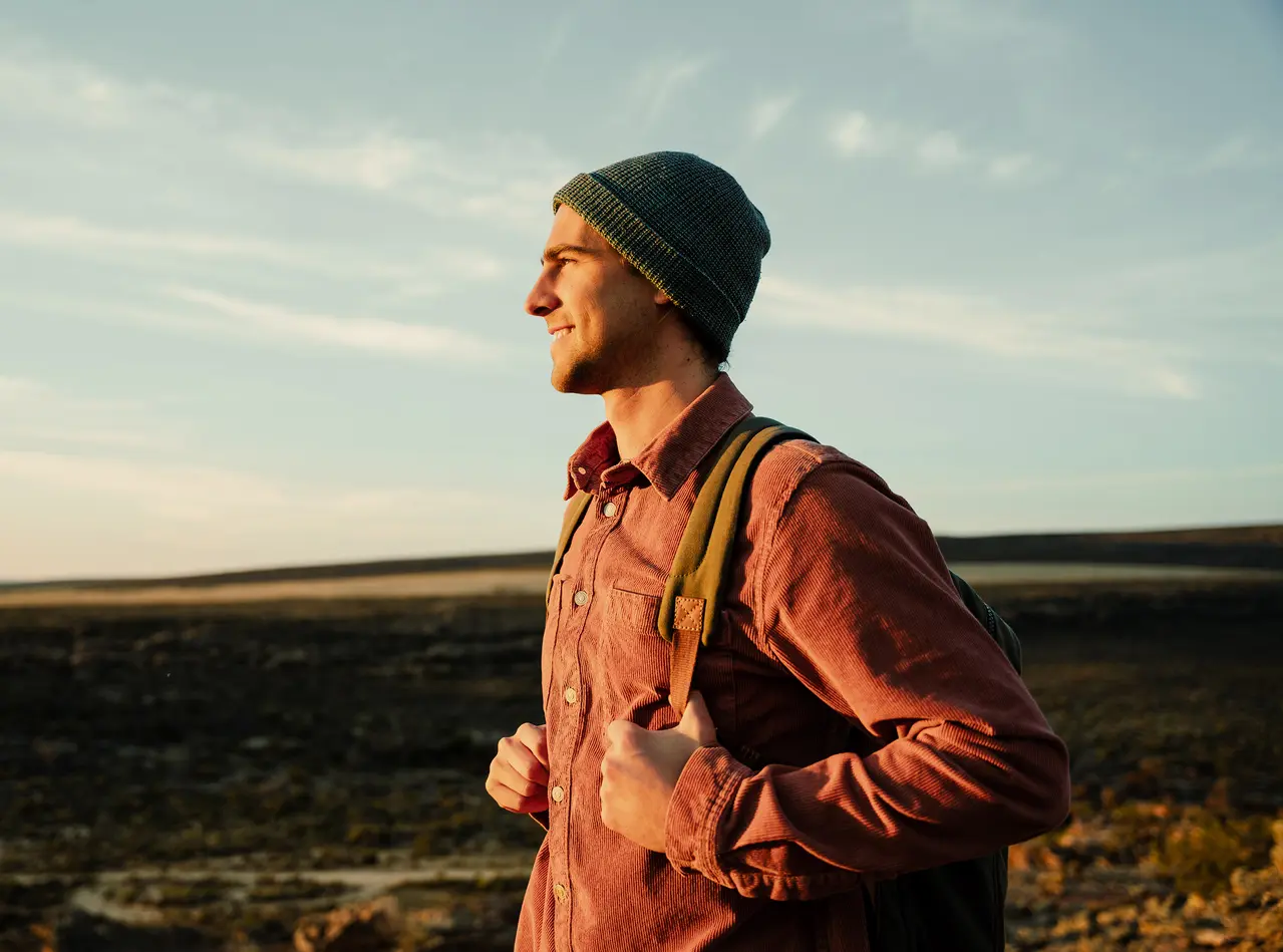 Ein Mann schaut mit einem lächeln Richtung Horizont. Er trägt eine Jacke, Mütze und einen Rucksack in der Natur.
