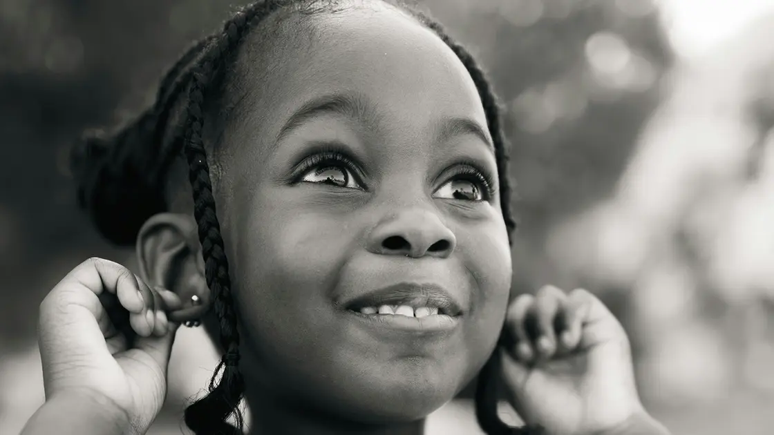 Ein lächelndes junges Mädchen mit geflochtenen Haaren blickt auf und hält ihre Hände an den Ohren, während sie im Freien steht.