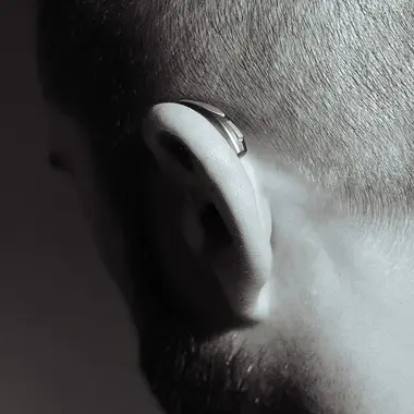 Eine Person mit rasiertem Kopf und Irokesenschnitt wird von hinten in Schwarz-Weiß gezeigt. Die Person trägt auch ein Hörgerät.