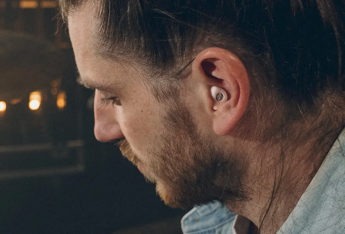 Mann mit kurzen braunen Haaren und Bart, der ein Hörgerät trägt, mit Blick nach unten.