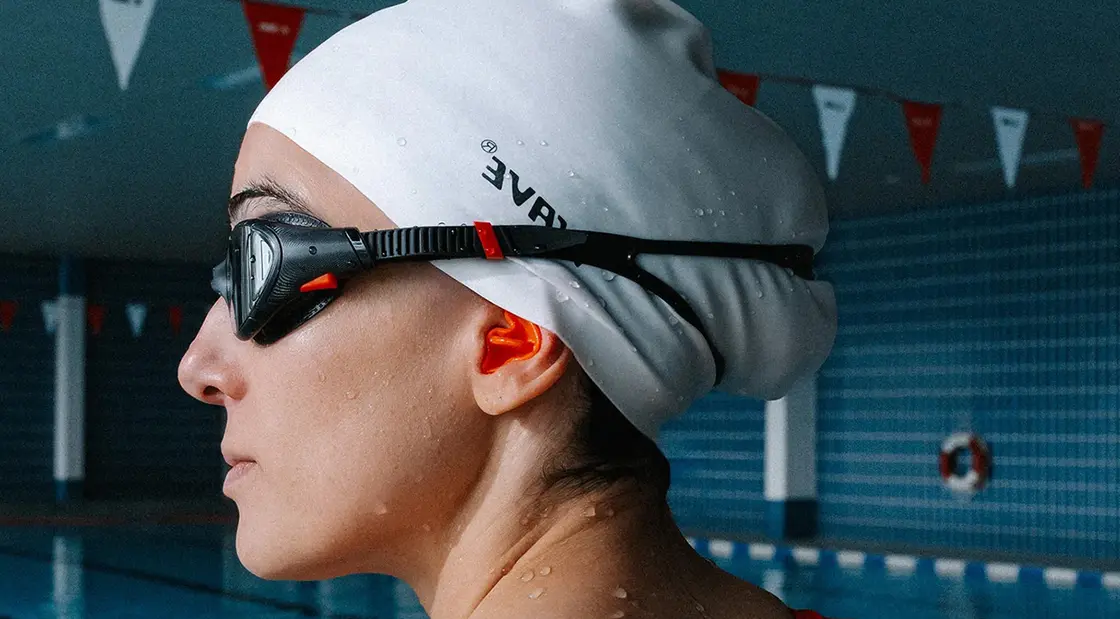 Nahaufnahme eines Schwimmers mit weißer Badekappe und schwarzer Schwimmbrille, der in einem Hallenbad steht.