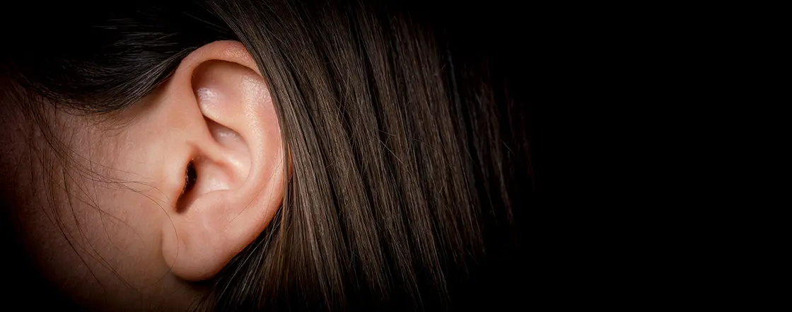 Nahaufnahme des Ohrs einer Person mit braunem Haar vor einem dunklen Hintergrund.