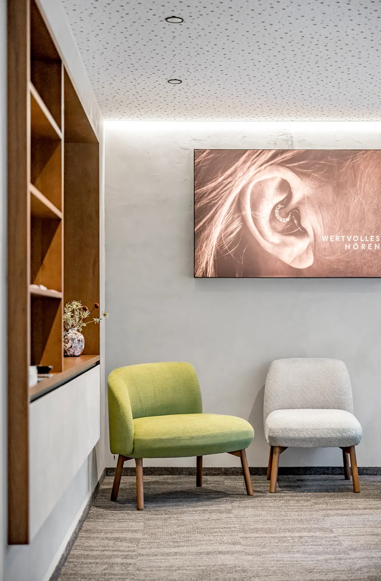 Zwei moderne Stühle, ein grüner und ein grauer, stehen neben einem Holzregal unter einem Bild in dem hellen, stilvollen Wartebereich.