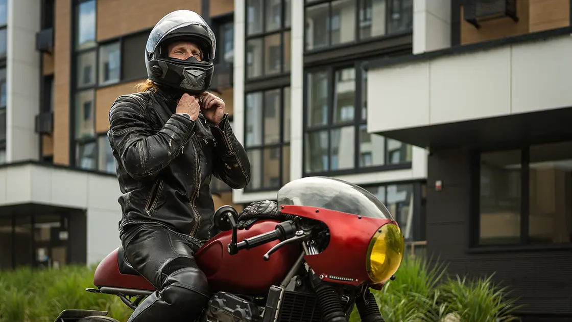 Eine Person in schwarzer Motorradkleidung passt ihren Helm an, während sie auf einem roten Motorrad vor einem modernen Gebäude sitzt.