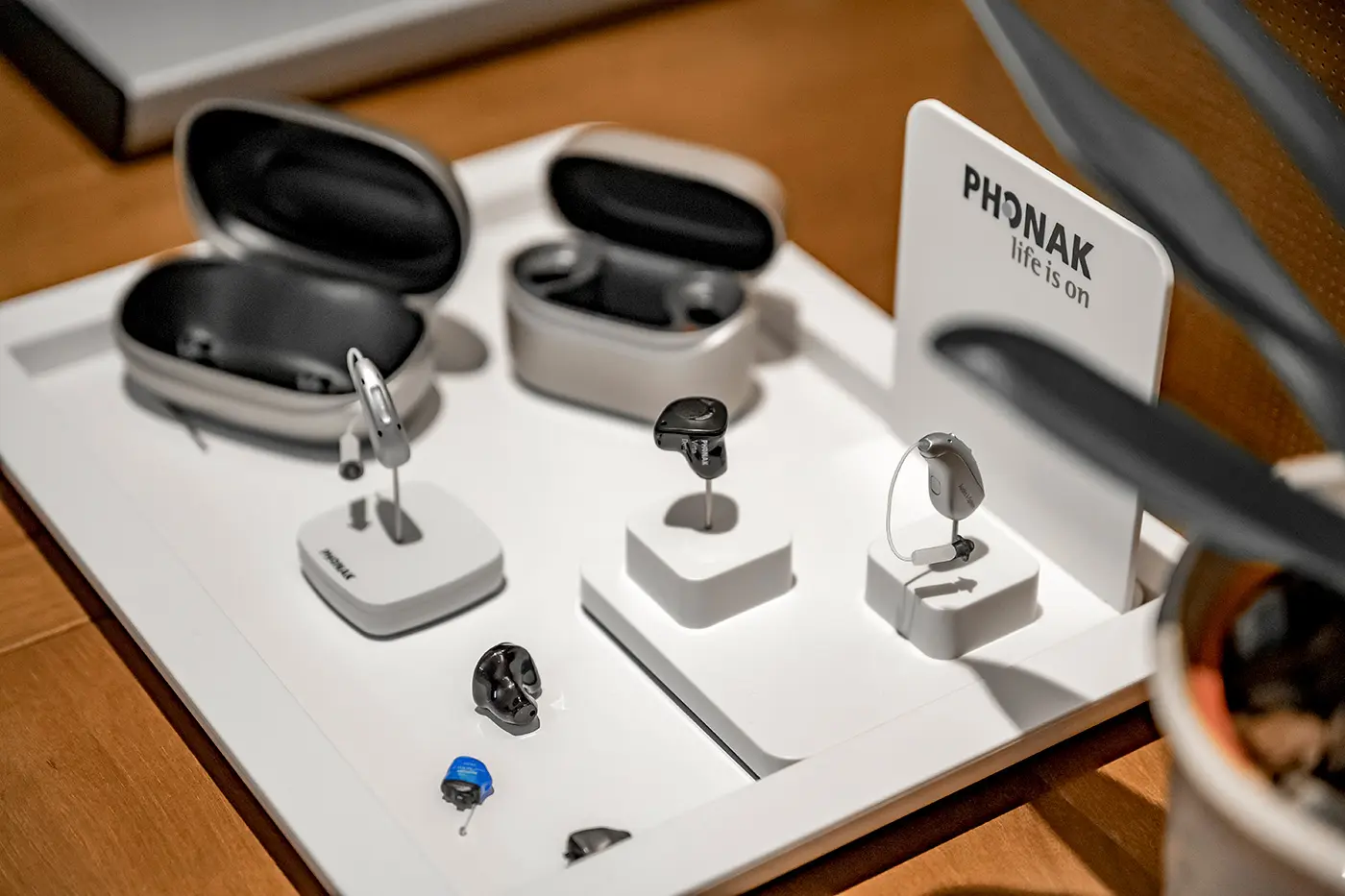 Ein Schautablett mit verschiedenen Hörgeräten, Ohrstöpseln und offenen Etuis auf einem Holztisch.