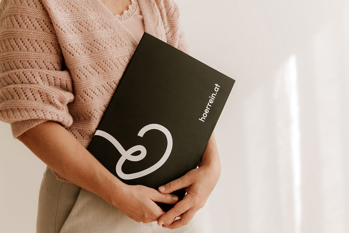 Eine Frau in einem rosa Pullover hält eine schwarze Mappe mit dem Hörrein Logo vor einem hellen Hintergrund.