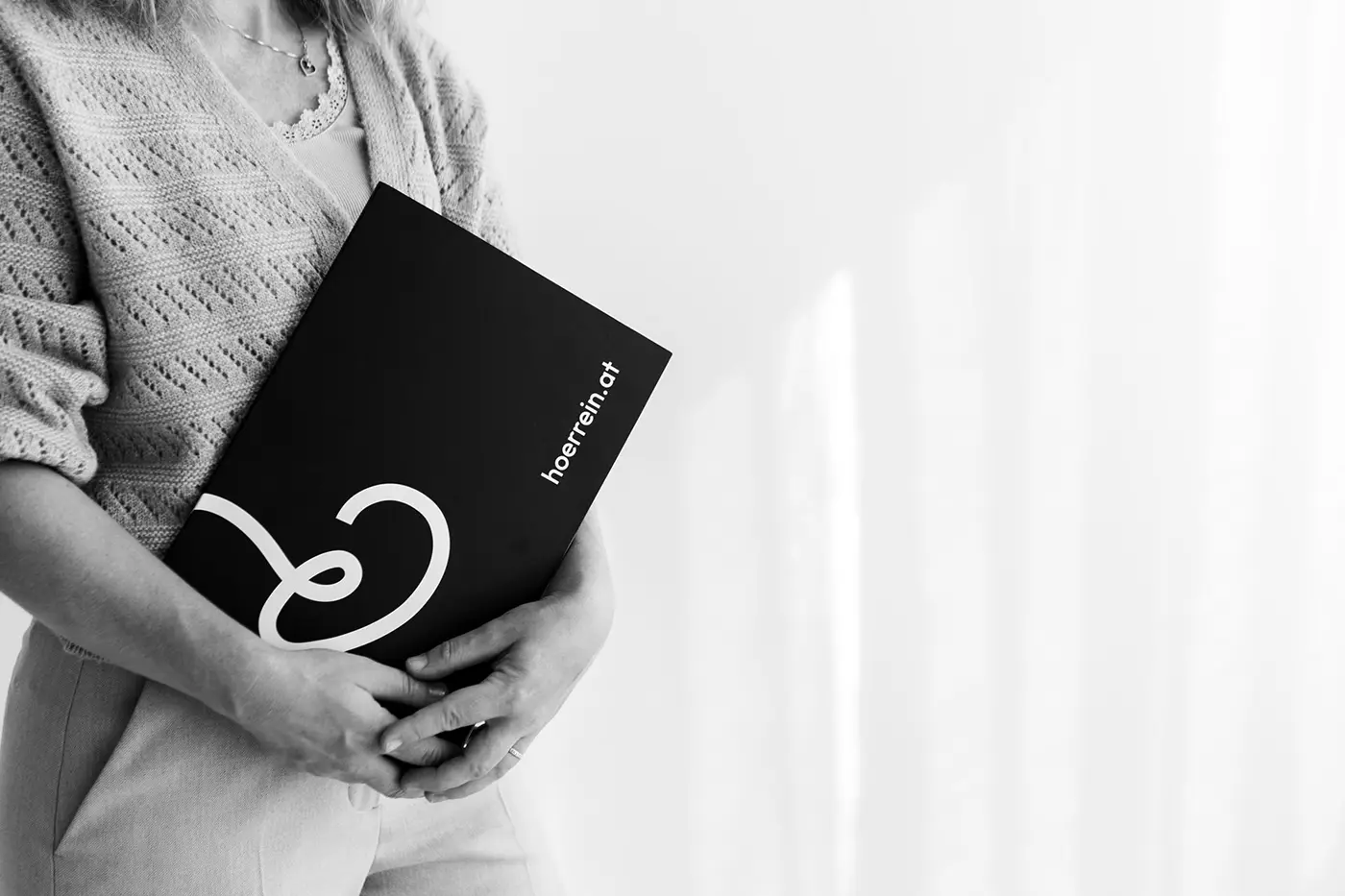 Schwarz-weiß Foto einer Frau in einem rosa Pullover, die eine schwarze Mappe mit dem Hörrein Logo vor einem hellen Hintergrund hält.