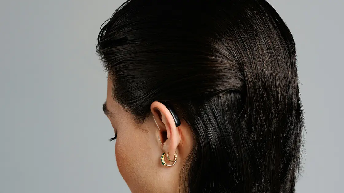 Frau mit zurückgekämmten dunklen Haaren, die kleine Ohrringe und ein Hörgerät trägt, von hinten vor einem schlichten Hintergrund gezeigt.