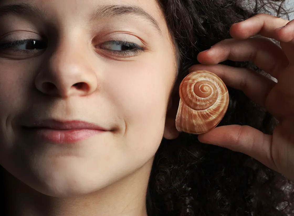 Lächelndes Kind, das sich eine Muschel ans Ohr hält und sie anschaut.