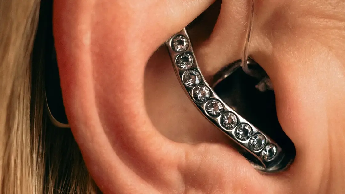 Nahaufnahme eines Otoplastik-Hörgerätes. Es passt sich dem Ohr der Dame genau an und ist mit Strasssteinen versetzt.
