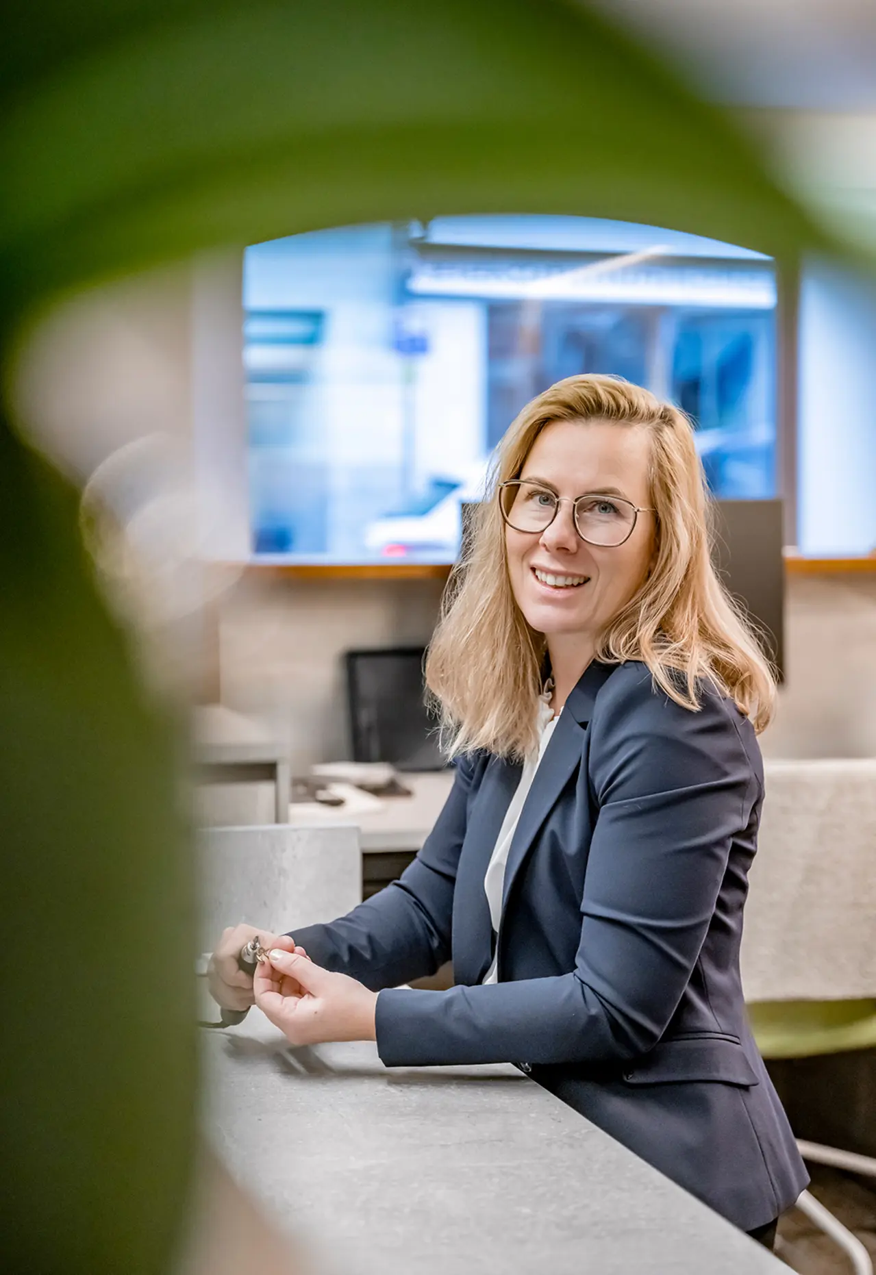 Eine Frau mit blondem Haar und Brille sitzt lächelnd an einem Schreibtisch in einem modernen Büro, teilweise eingerahmt von einer Pflanze.