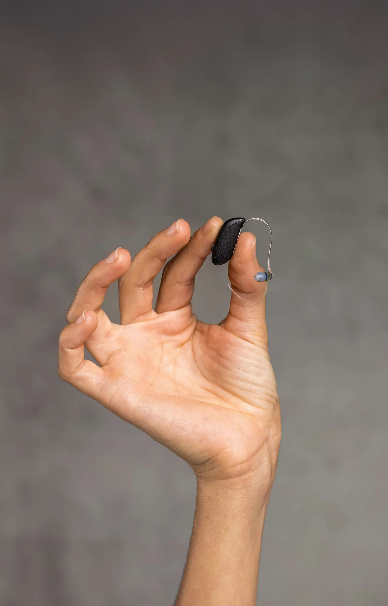 Es ist ein Arm zu sehen, der ein Hörgerät in die Luft hält. Schlichter Hintergrund.