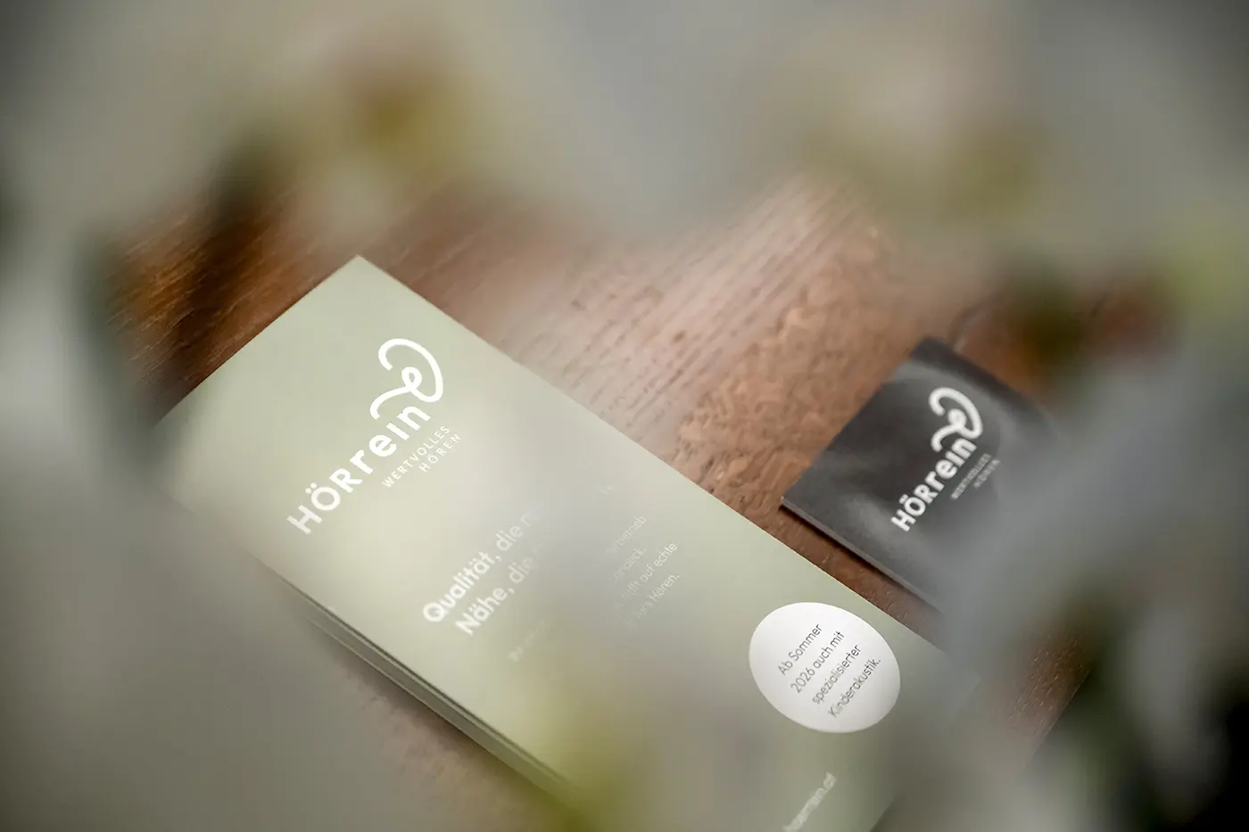 Grüne Flyer und Visitenkarten auf einer hölzernen Oberfläche, teilweise verdeckt durch eine verschwommene grüne Pflanze.