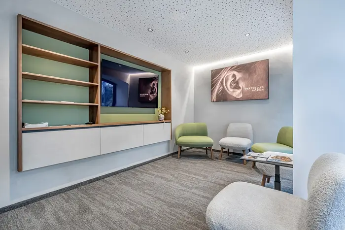 Modernes Wartezimmer mit grünen Stühlen, weißer, strukturierter Decke, Wandkunst und eingebauten Holzregalen an der linken Wand.