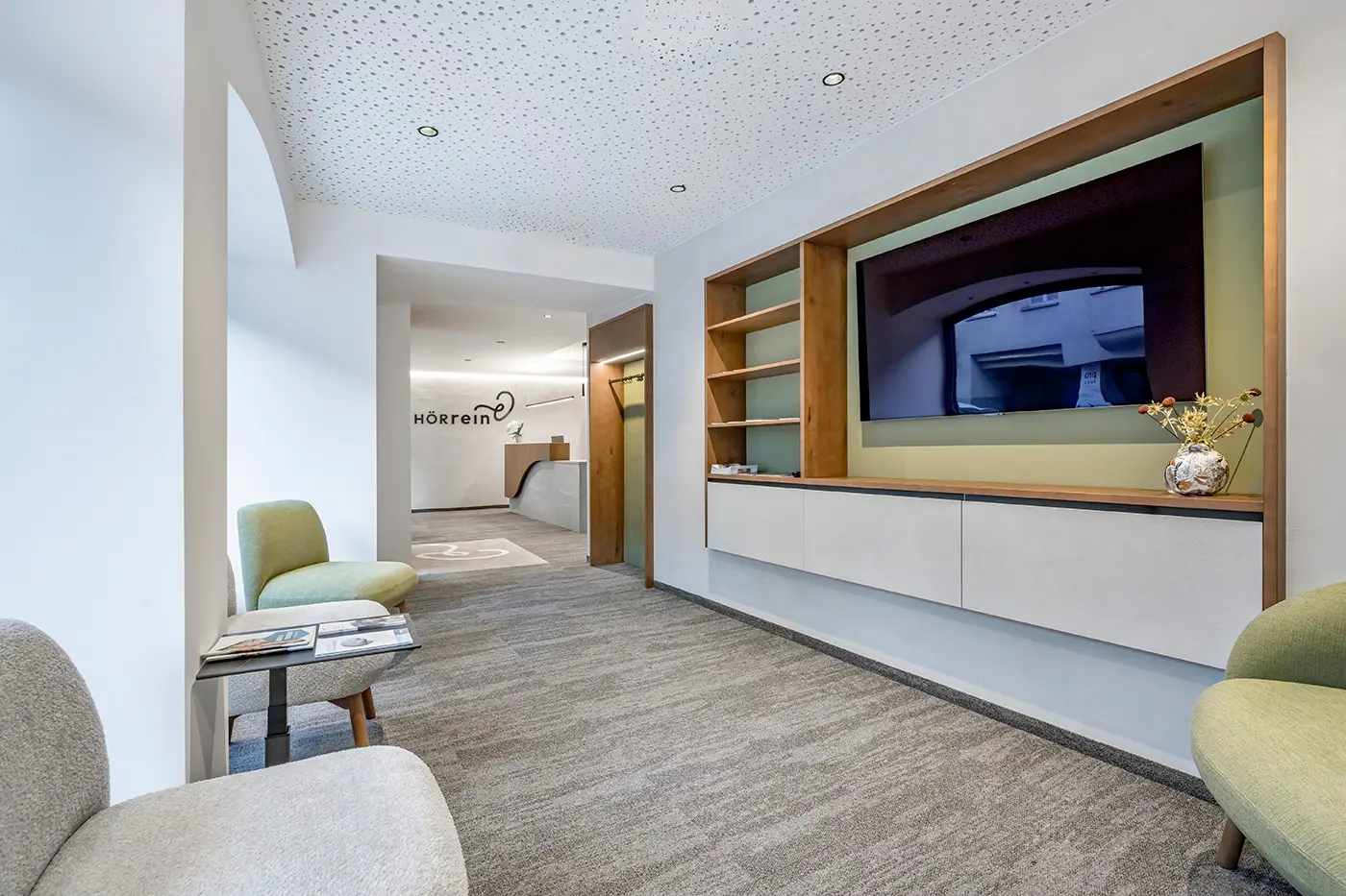 Modernes Wartezimmer mit Sesseln, einem Fernseher in einer Schrankwand und einem strukturierten Teppichboden mit Blick zur Rezeption.