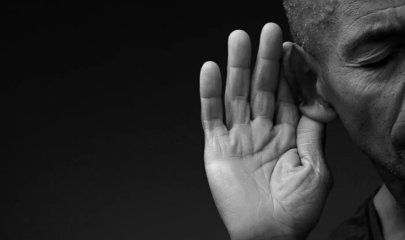 Eine Person hält sich vor einem dunklen Hintergrund die Hand ans Ohr, als würde sie genau zuhören.