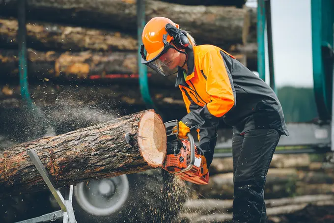 Eine Person in Schutzausrüstung sägt mit einer Kettensäge einen Baumstamm, während im Hintergrund Sägespäne aufgewirbelt werden und Stämme gestapelt werden.