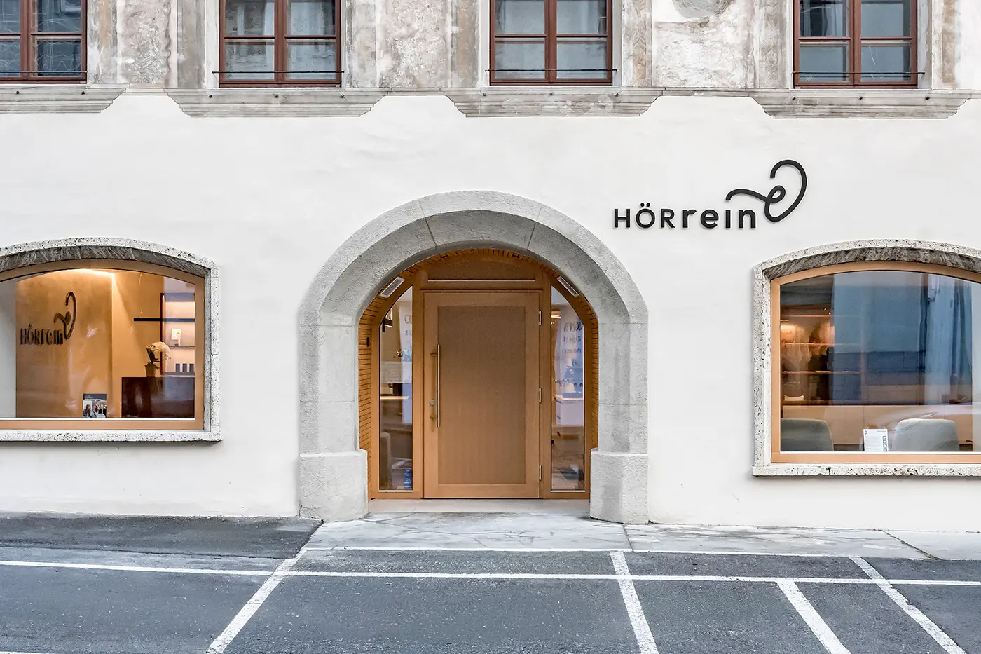 Fassade mit gewölbter Holztür und großen Fenstern mit dem HÖRrein Logo über dem Eingang.