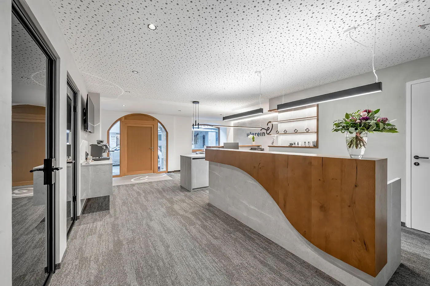 Moderner Empfangsbereich mit einem geschwungenen Holzschreibtisch, grauem Teppich, weißen Wänden und einer Blumenvase auf dem Tresen.