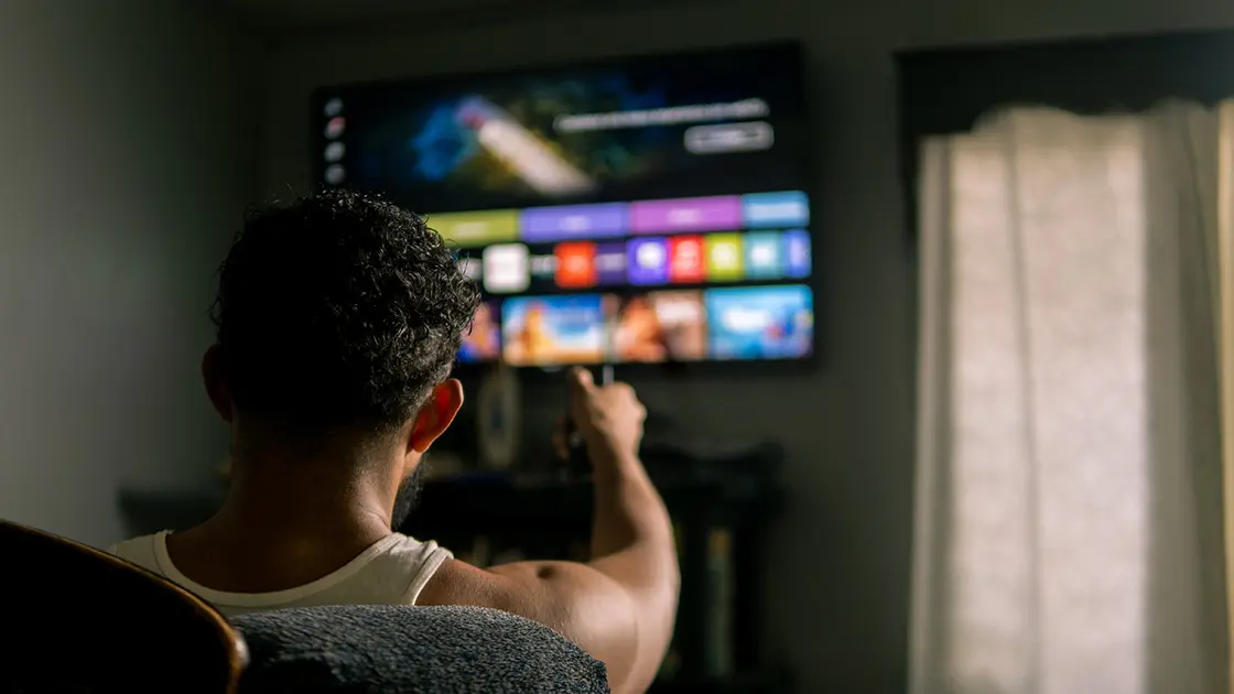 Eine Person mit kurzen lockigen Haaren zeigt mit der Fernbedienung auf einen Smart-TV, der in einem schwach beleuchteten Raum Streaming-Apps anzeigt.