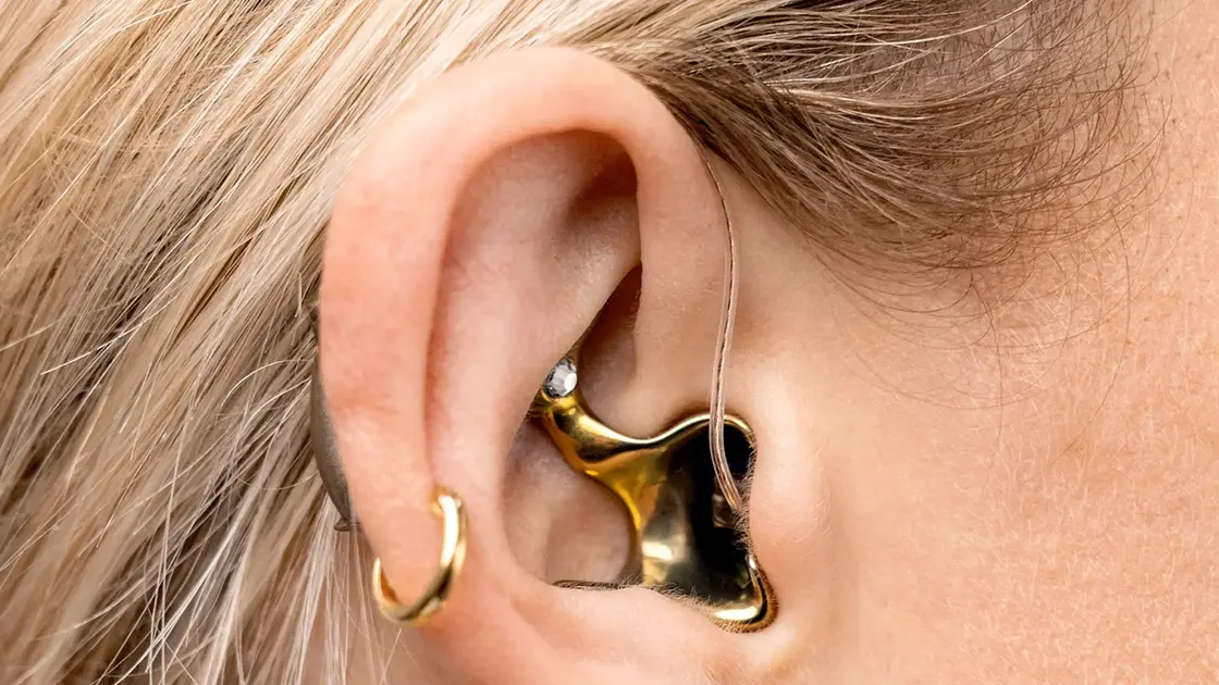 Nahaufnahme eines Ohrs mit mehreren goldenen Ohrringen und einem goldglänzenden Otoplastik Hörgerät..