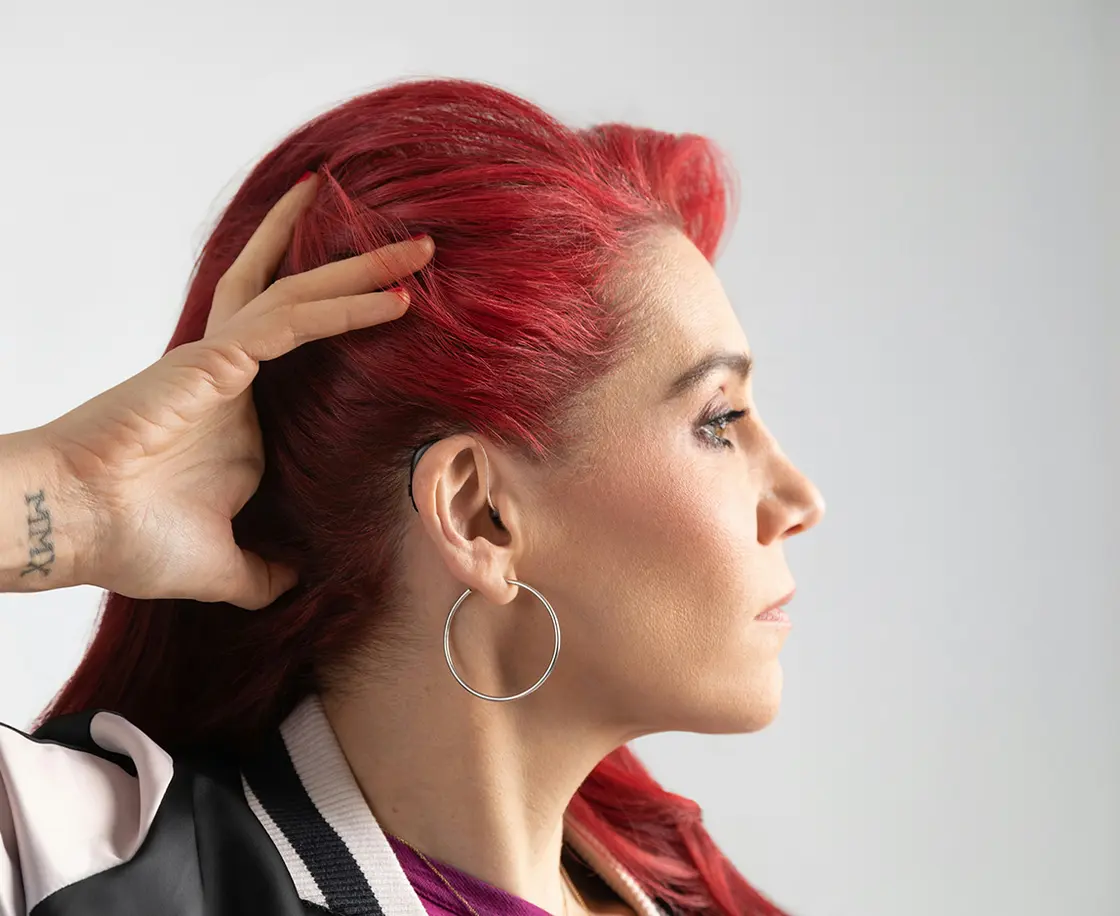 Person mit langen roten Haaren im Seitenprofil, die Hand berührt den Kopf, sie trägt Hörgeräte und eine leichte Jacke.