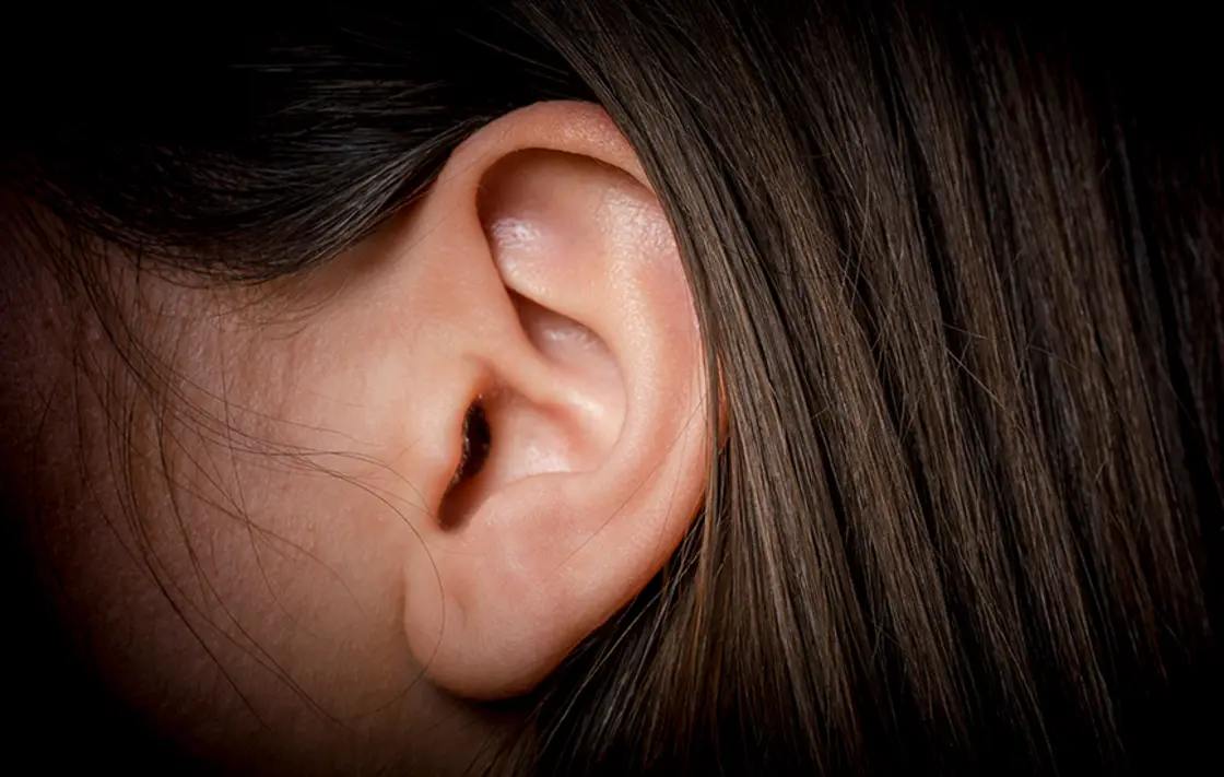 Nahaufnahme des Ohrs einer Person mit braunem Haar vor einem dunklen Hintergrund.