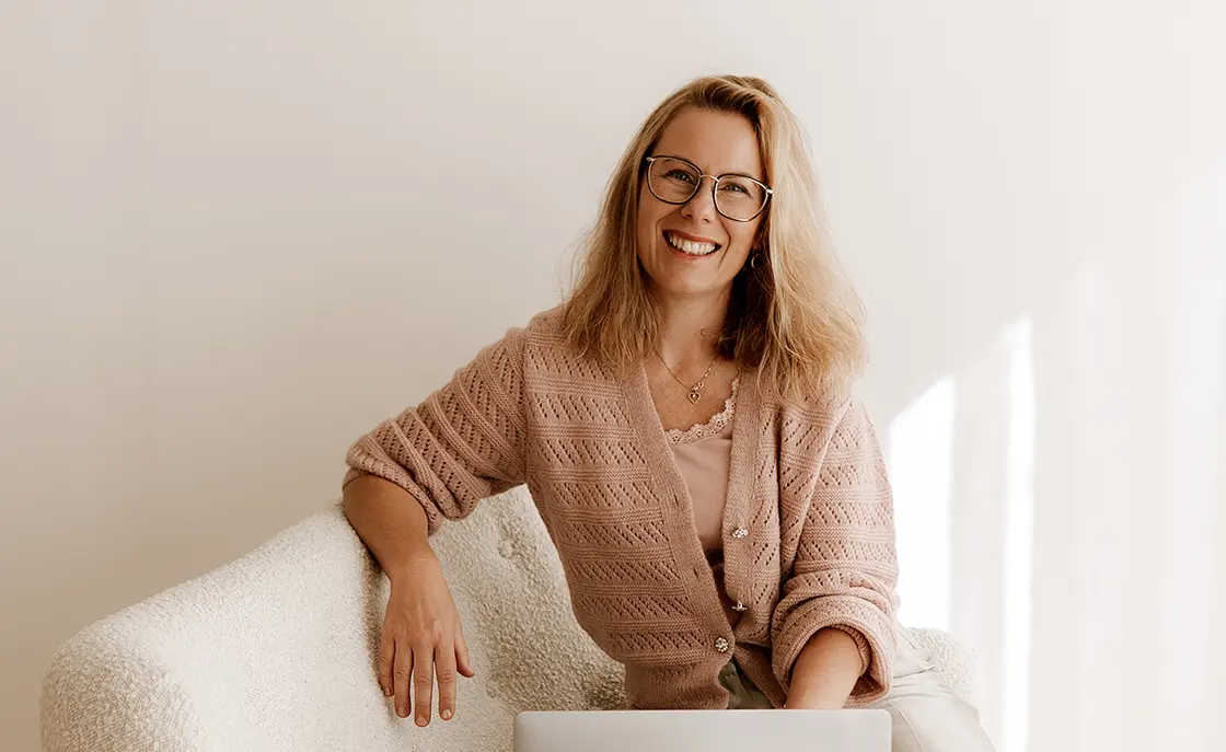 Geschäftsführerin sitzt mit ihrem Laptop auf einem weißen Sofa und lächelt in die Kamera. Sie hat schulterlange blonde Haare und ein pinkes Oberteil an.