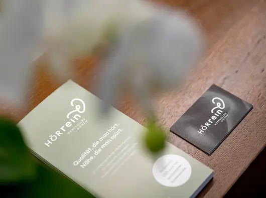 Eine grüne Broschüre und schwarzen Visitenkarten mit weißem Text und Logos auf einer hölzernen Oberfläche, die teilweise durch eine grüne Pflanze verwischt wird.