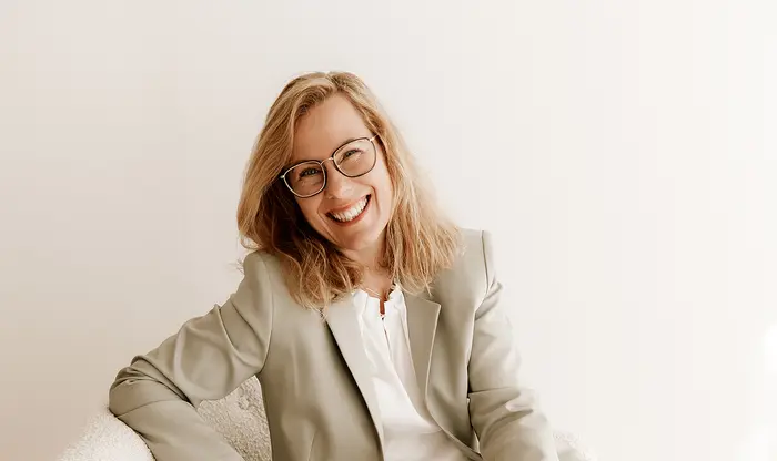 Die Geschäftsführerin Janett Schöpf mit Brille und lockigem Haar, die einen hellen Blazer und ein weißes Hemd trägt und vor einem schlichten Hintergrund sitzt.