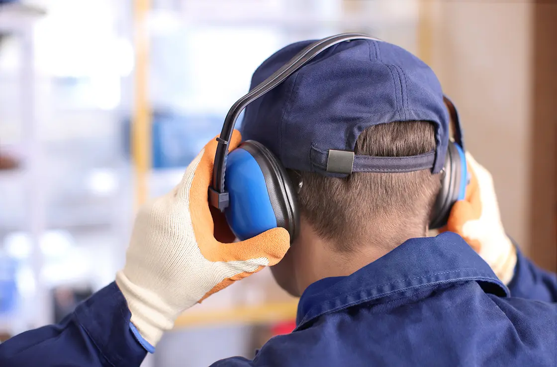 Eine Person mit Schutzhandschuhen und Mütze setzt sich in einer Industrieumgebung einen blauen Gehörschutzkopfhörer auf.
