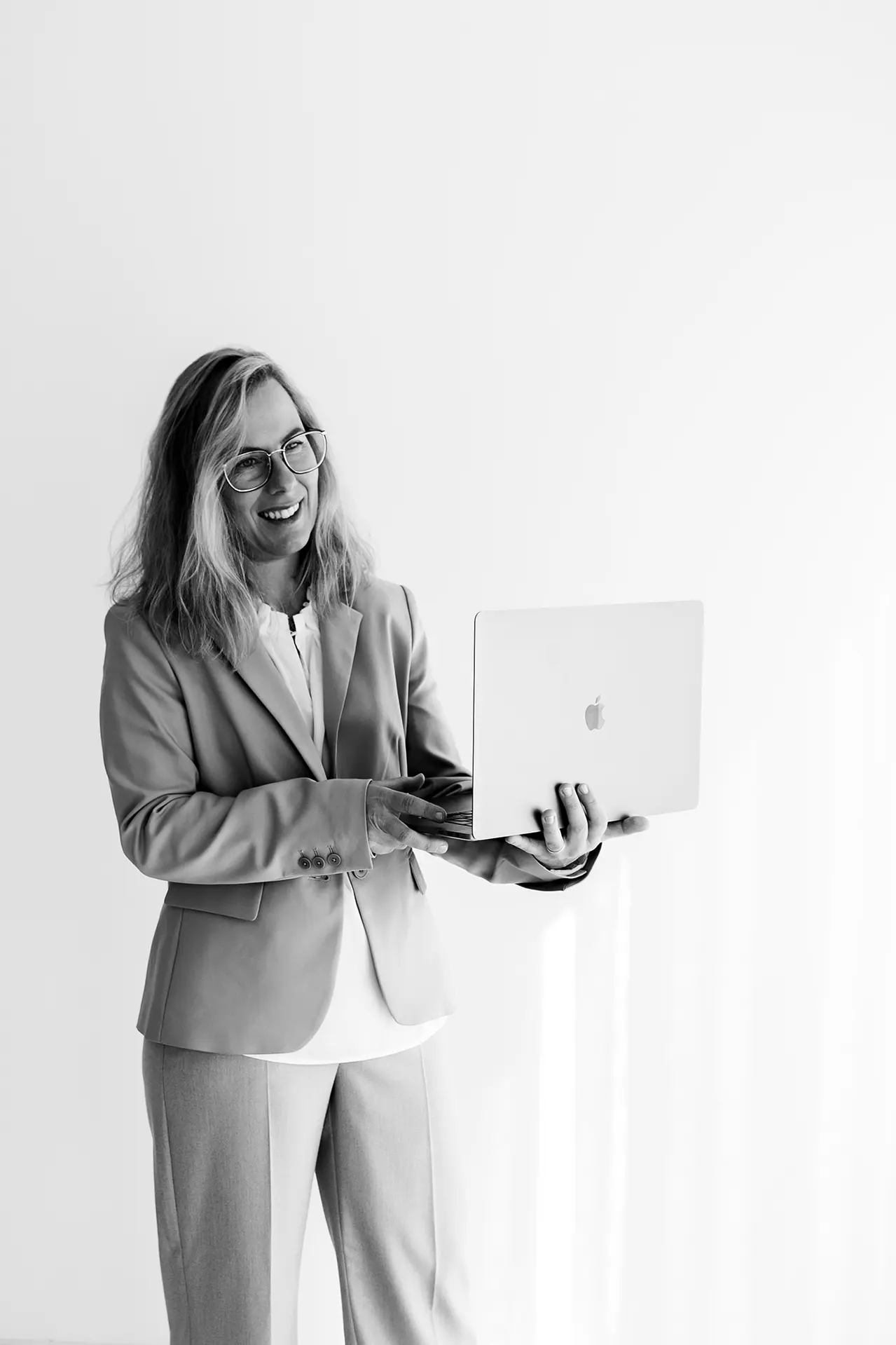 Ein schwarz-weiß Foto der Geschäftsführerin im Anzug. Sie steht im Raum und hält lächelnd ihren Laptop in den Händen.