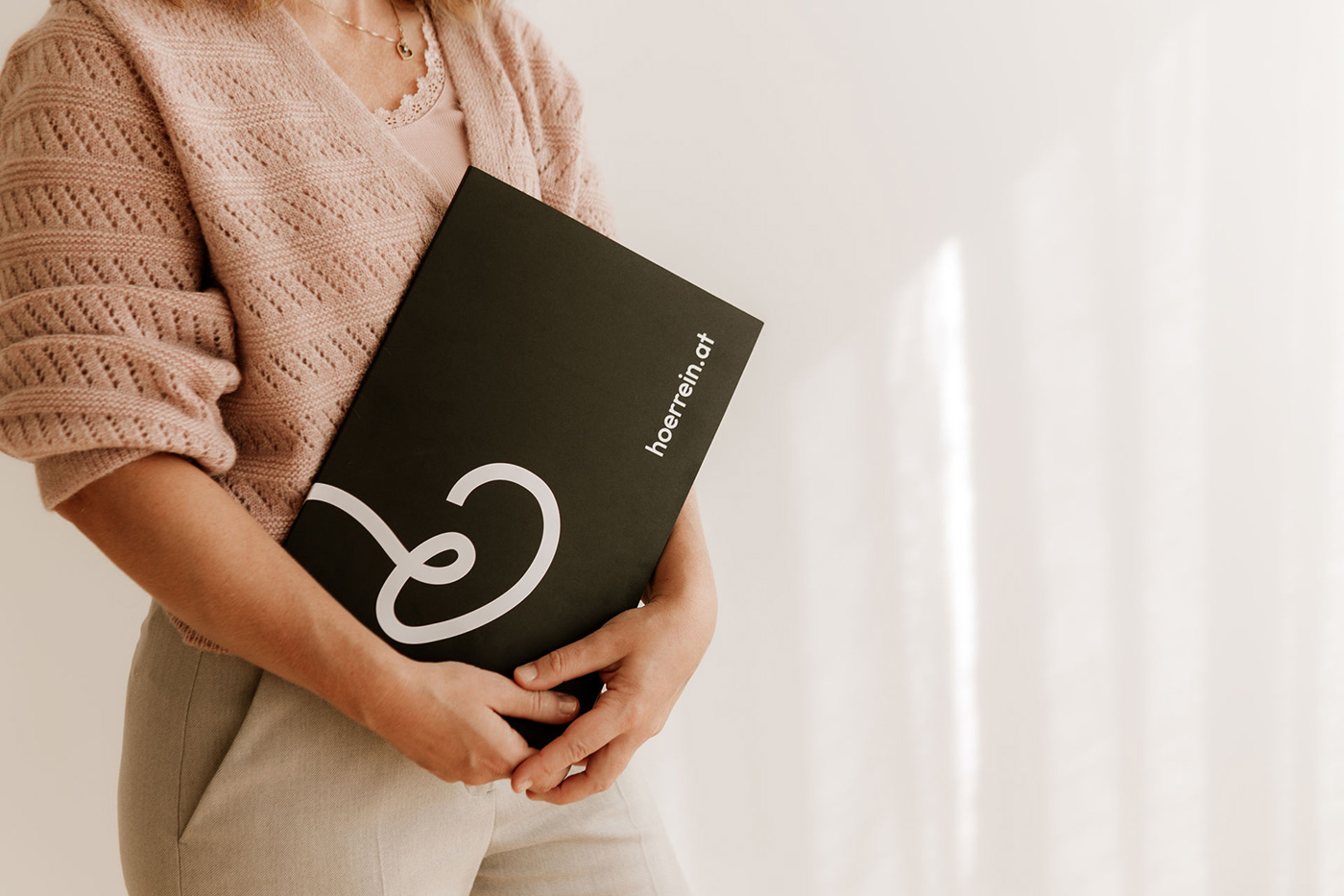 Eine Frau in einem rosa Pullover hält eine schwarze Mappe mit dem Hörrein Logo vor einem hellen Hintergrund.